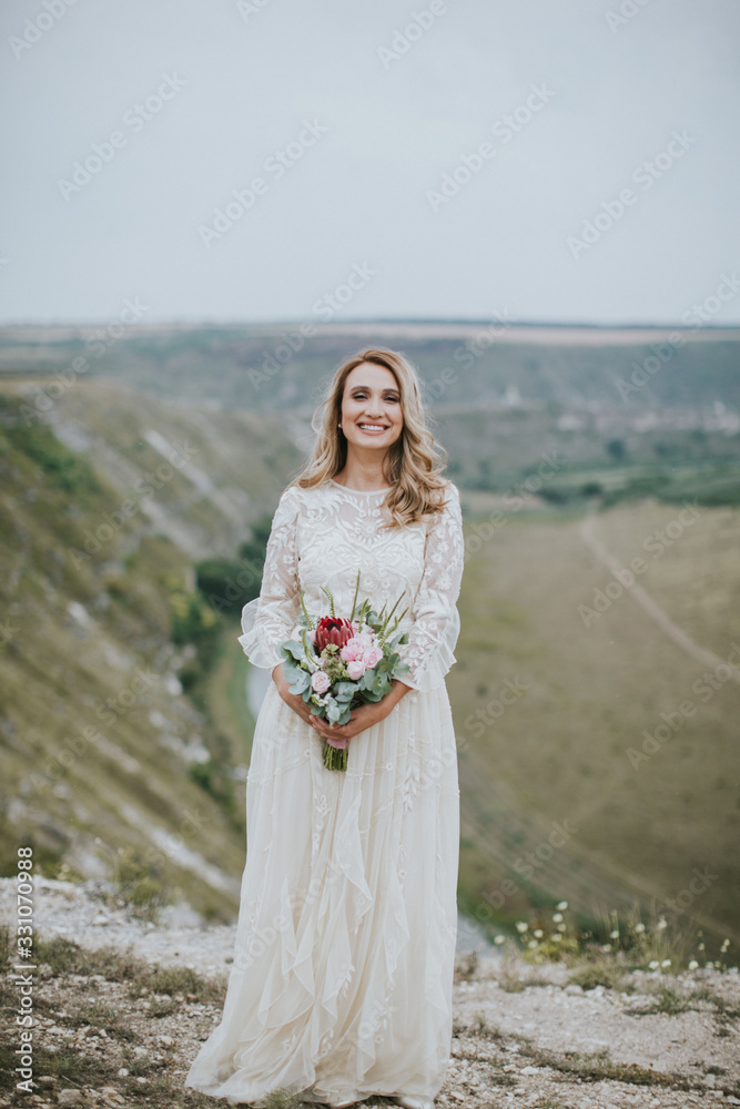 Smiling young blonde caucasian bride in a white, modern lightweight dress posing. Wedding bouquet in her hands. Happy vintage hipster bride, warm colors.