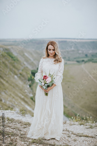 Smiling young blonde caucasian bride in a white, modern lightweight dress posing. Wedding bouquet in her hands. Happy vintage hipster bride, warm colors.