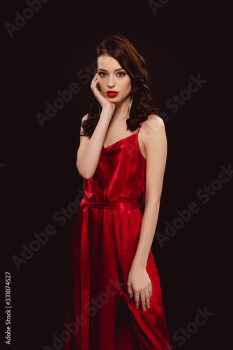 Beautiful woman in red dress touching face and looking at camera isolated on black