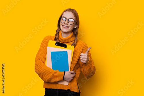 Happy red haired caucasian student with freckles and eyeglasses is holding some books and gesturing the like sign on a yellow background