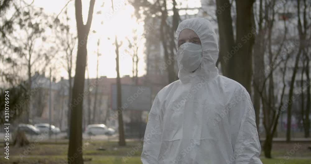 Portrait of male scientist in protective suit adjusting his mask in daytime park in pandemic coronavirus quarantine time as cars and pedestrians pass by 4K Portrait of male scientist in protective suit adjusting his mask in daytime park in pandemic coronavirus quarantine time as cars and pedestrians pass by 4K