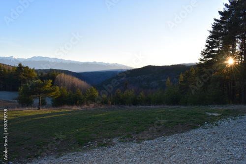 Sunbeams through evergreen pine trees in southeast European mountain resort View from an amusement park attached to a bear sanctuary with alpine coaster in Belitsa village A snow covered peaks horizon photo