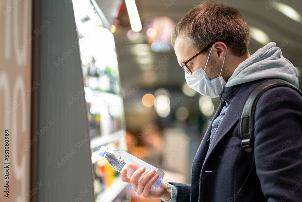 Caucasian man customer in glasses wearing facial protective mask, buying and choosing water bottle, reads information on a label. Purchase of food during the coronavirus, covid-19 epidemic in USA.  - obrazy, fototapety, plakaty 