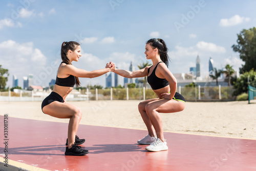 Strength in teamwork. Two young attractive women athletes exercise on the beach doing squats with a sunrise and ocean in the background.