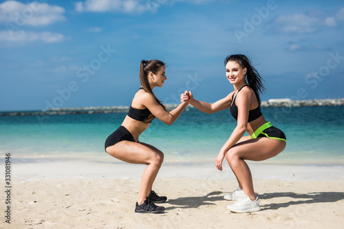 Two young fitness women doing squats exercise on beach