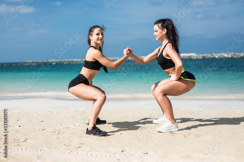 Strength in teamwork. Two young attractive women athletes exercise doing squats on the beach near beach