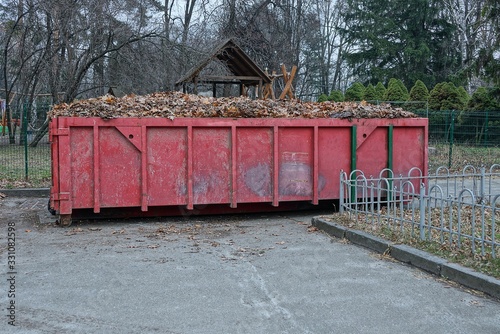 one big red iron dumpster full of dry leaves stands on the pavement in a park