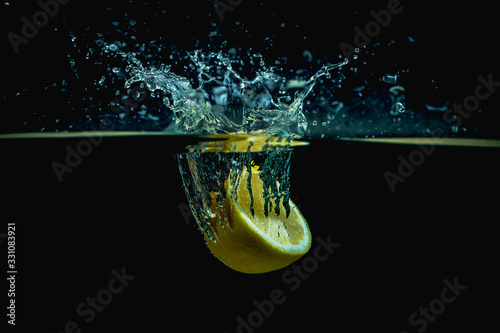 Fresh lemon half falls into water making big splash of drops and spray on a dark background. Juicy and fresh concept of lemon in water making a beautiful water crown.