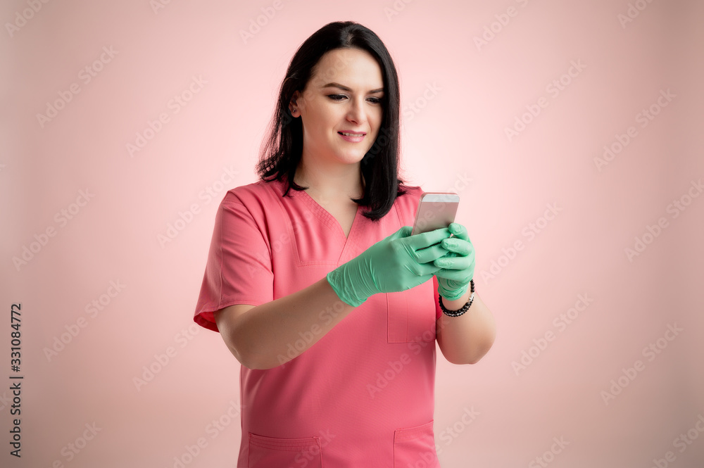 Beautiful woman doctor with stethoscope, wearing pink scrubs holding smartphone, texting messages happy face Beautiful woman doctor with stethoscope, wearing pink scrubs holding smartphone, texting messages happy face