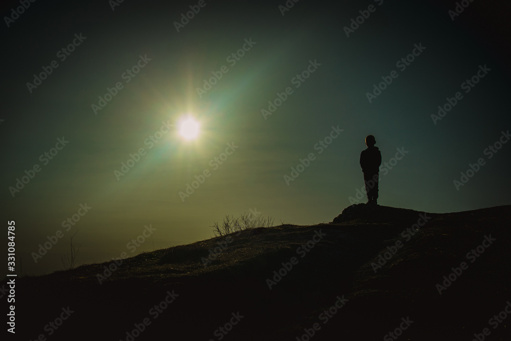 Silhouette of a young kid standing still on the edge of a hill or mountain with a magnificent sky backdrop behind him with sun some lens flare.