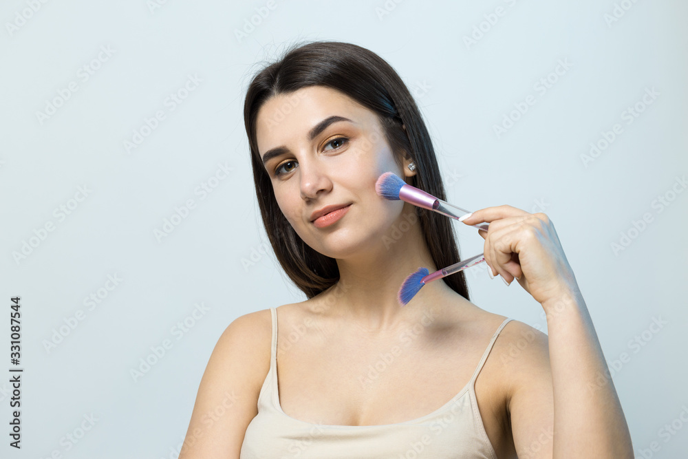 Close-up of a young girl in a light top on a white background making a facial make-up. A pretty woman holds a cosmetic brush near her face and smiles.