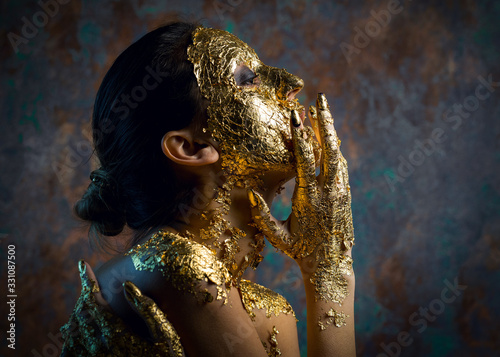 Girl with a mask on her face made of gold leaf. Gloomy studio portrait of a brunette on an abstract background.