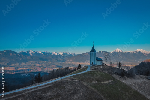 Aerial drone photo of cute fantastic charming Saint Primoz church on a small hill with mountains in background at sunset, Jamnik village, Slovenia, Europe with picturesque church in early spring.