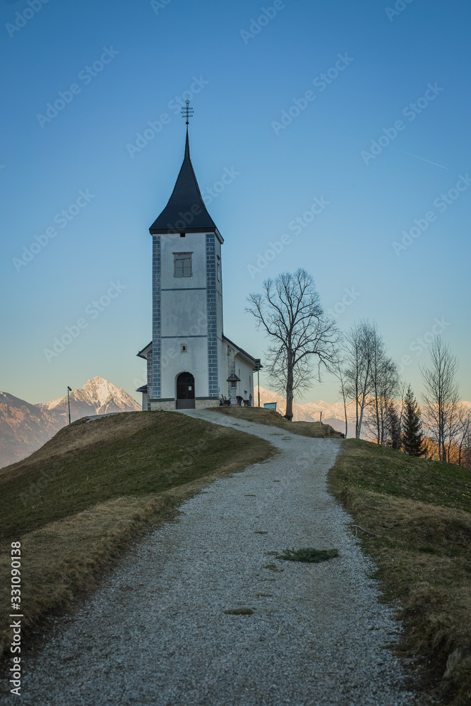 Path towards cute  fantastic charming Saint Primoz church on a small hill with mountains in background at sunset, Jamnik village, Slovenia, Europe with picturesque church in early spring.
