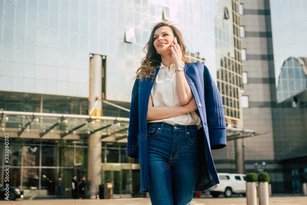 Caucasian business woman speaking by phone. Waist up portrait of a successful European business woman woman, talking on the phone, standing on glass background, modern office building. Sunny weather