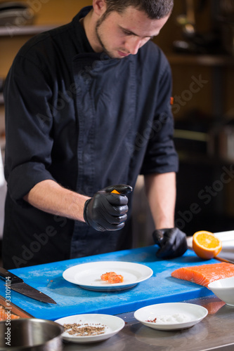 Theme cooking is profession of cooking. Portrait of Caucasian man in restaurant kitchen preparing red fish fillets salmon meat in black gloves uniform. Chef holds plate with blue and checks  tastes