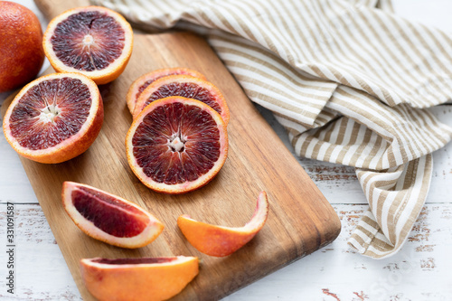 Close Up of Blood Oranges on a Wooden Cutting Board on a White Tabletop; Kitchen Towel Beside Cutting Board; One Whole; One Cut in Half; Some Slices and Wedges