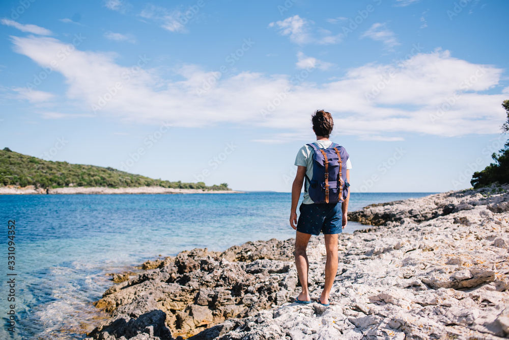 Back view of male traveler with backpack looking at beautiful seascape and calm water surface exploring nature, young 20s man in casual wear enjoying free time and getting to destination on castline