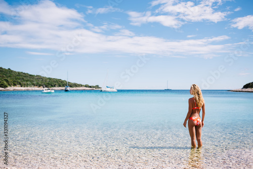 Back view of blonde female 20s traveler relaxing on coachtline on seaside in marine harbor recreating on vacations, young woman in swimsuit sunbathing and enjoying warm weather on ocean near shore
