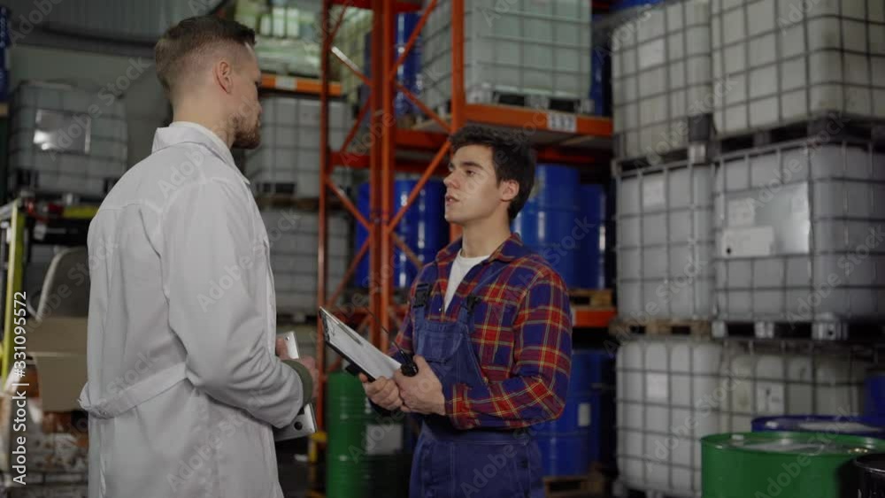 Young industrial warehouse worker meeting engineer in white coat and showing him report on clipboard in factory storage Young industrial warehouse worker meeting engineer in white coat and showing him report on clipboard in factory storage