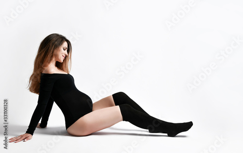 Pregnant woman in black leotard and stockings. She looking at her tummy and smiling, sitting on floor, posing isolated on white.