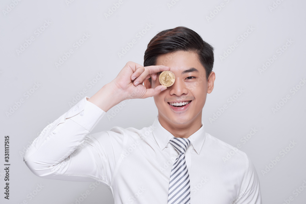 Young man standing isolated on white background holding cryptocurrency coin covering eye looking aside smiling playful