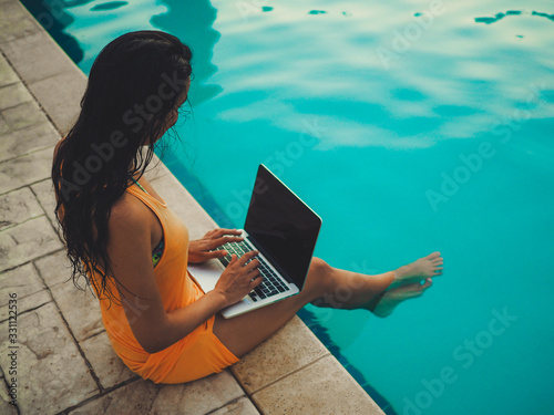 remote online working digital nomad women from the side with long hair and laptop sitting at a sunny blue water pool  photo