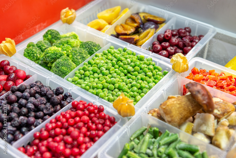 Frozen fruits and vegetables. Berries, mushrooms, beans and more. Products are poured into plastic boxes. In the center of the frame is green peas.