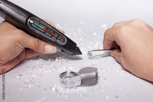 Diamonds on tweezers with testing machine on a white background, reflections on the ground. brilliant cut diamond held by right hand, tester, magnifier , diamond checking equipment.