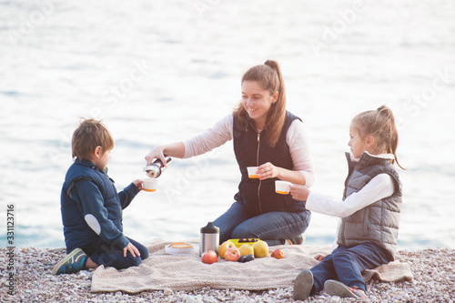 happy family mother and two kids in spring clothes drink hot tea on windy cold sea beach during leisure picnic