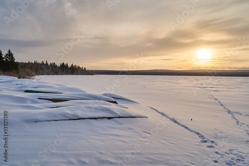     Snow- covered lake at dawn with boats on the shore.  Winter forest with snow-covered fir trees high in the mountains. Sunny February day in the spruce forest.      