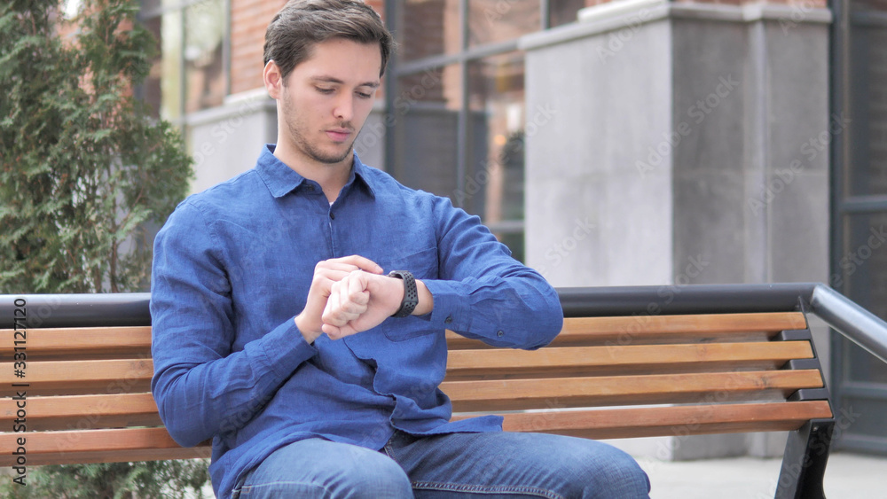 Fototapeta premium Young Man Using Smartwatch, Outdoor Sitting on bench