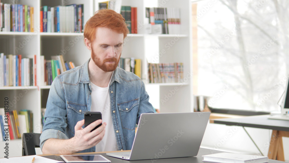 Young Casual Redhead Man Using Phone and Laptop Young Casual Redhead Man Using Phone and Laptop