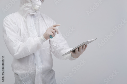 man wearing biological protective uniform suit clothing, mask, gloves spraying sanitizer on tablet for sanitizing virus bacteria