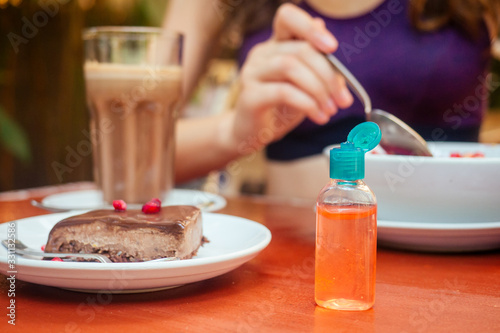 woman useing antibacterial gel on hands before start to eating her healthy food in tropical asian cafe . smoothie bowl , raw cake and coffee on table with sanitizer on bottle