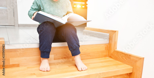 The child is reading the paper book sitting on the wooden stairs in the house