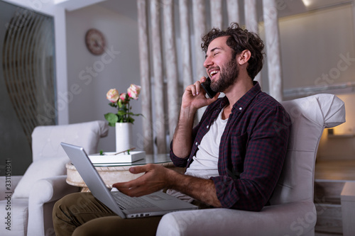 Young attractive smiling guy is browsing at his laptop and talking oh mobile phone, sitting at home on the cozy sofa, wearing casual outfit. Freelance business work from home concept