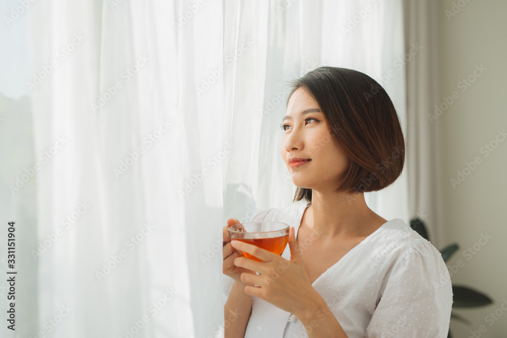 Portrait of a satisfied woman thinking looking at side holding a tea mug beside a window at home Portrait of a satisfied woman thinking looking at side holding a tea mug beside a window at home