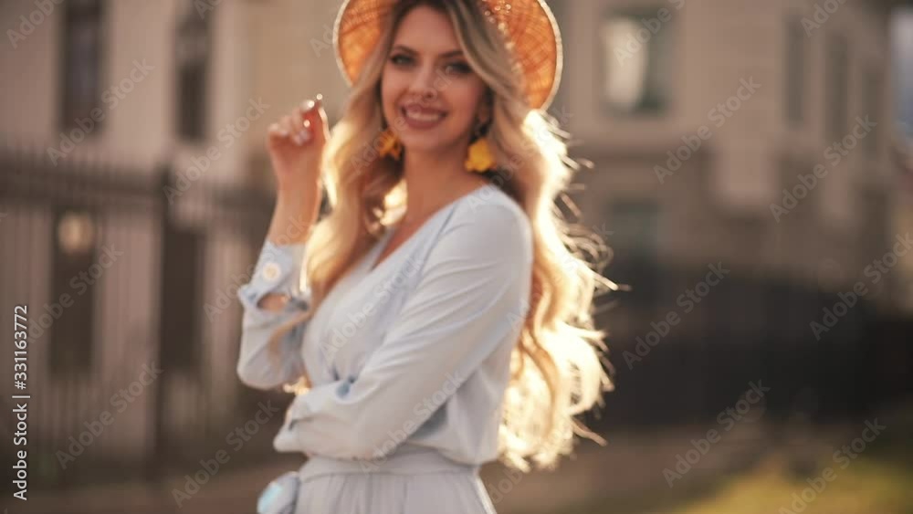 Stock video of a gorgeous blonde woman with long wavy hair wearing summer wattled hat and flower earrings posing to the camera in beautiful bright sunshine in the street. She is smiling at camera. Stock video of a gorgeous blonde woman with long wavy hair wearing summer wattled hat and flower earrings posing to the camera in beautiful bright sunshine in the street. She is smiling at camera.