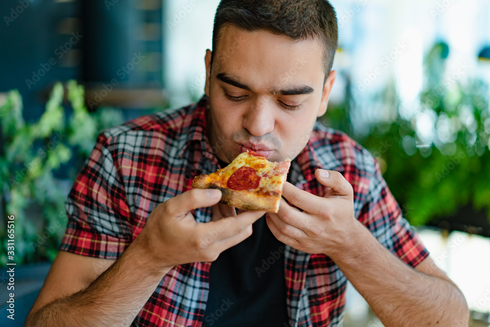 Handsome man in a red plaid shirt eats pizza at a cafe. Hungry man eats a slice of pizza with cheese, sausage and sauce. Close-up Handsome man in a red plaid shirt eats pizza at a cafe. Hungry man eats a slice of pizza with cheese, sausage and sauce. Close-up