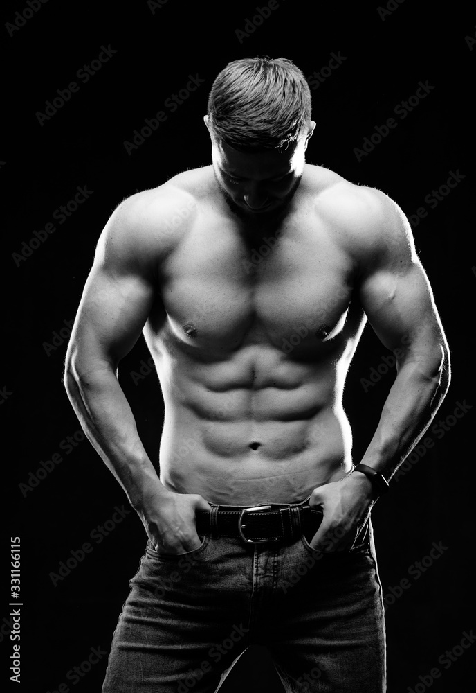 Muscular guy with naked torso posing with hands in pockets. Studio photo. Portrait of a handsome man in jeans on dark background. Black and white photo. Closeup. Muscular guy with naked torso posing with hands in pockets. Studio photo. Portrait of a handsome man in jeans on dark background. Black and white photo. Closeup.