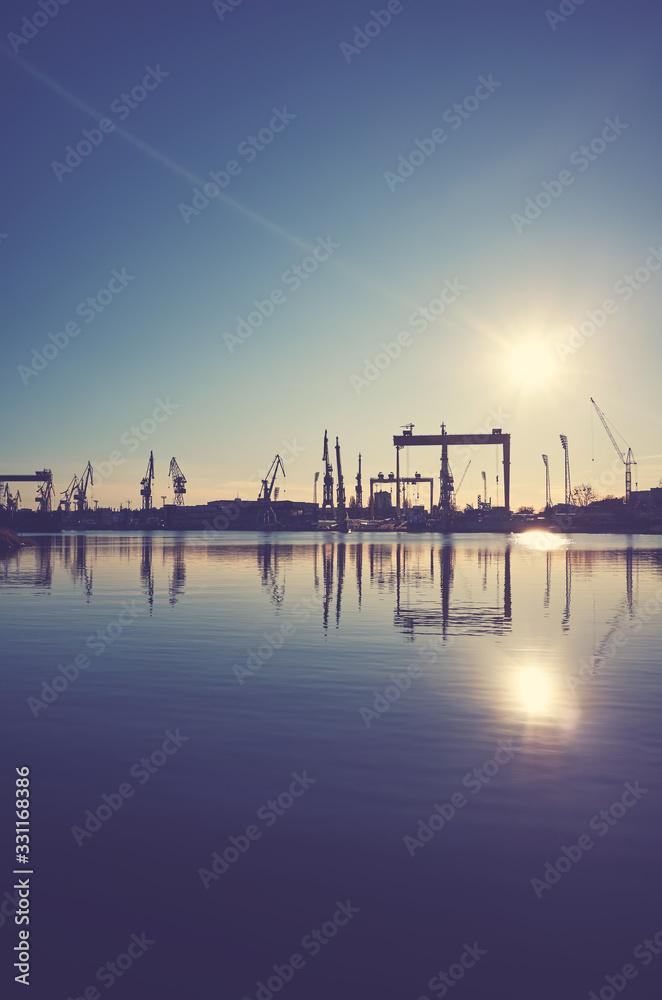 Shipyard cranes silhouettes reflected in water at sunset, color toning applied.