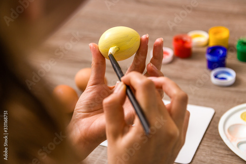 easter mood. close up young woman hands painting easter eggs with brush and gouache in pastel colors over wooden table at home