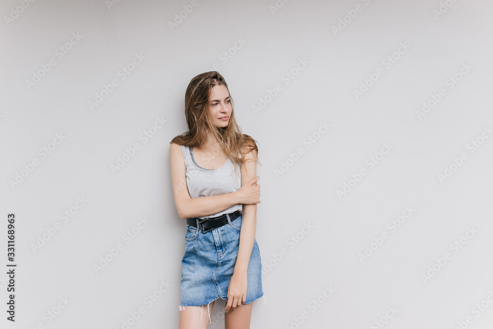Amazing woman in denim skirt posing in studio with white interior. Indoor shot of lovely brunette girl isolated on light background with pensive face expression. Amazing woman in denim skirt posing in studio with white interior. Indoor shot of lovely brunette girl isolated on light background with pensive face expression.