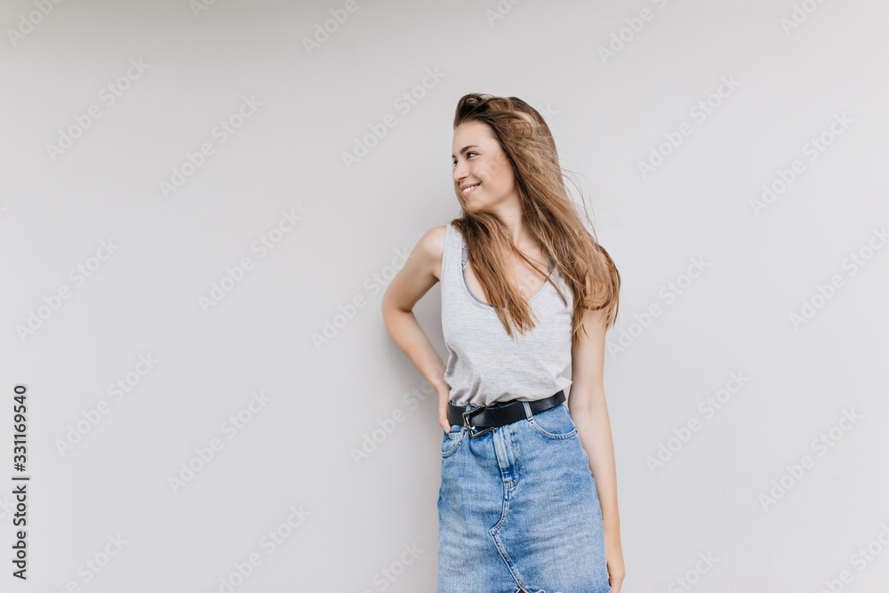 Ecstatic long-haired woman in stylish attire standing in confident pose in studio. Indoor portrait of inspired european girl wears trendy denim skirt. Ecstatic long-haired woman in stylish attire standing in confident pose in studio. Indoor portrait of inspired european girl wears trendy denim skirt.