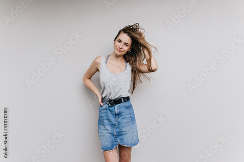Studio shot of charming woman in denim skirt posing with hair waving. Indoor photo of slim trendy girl isolated on light background.