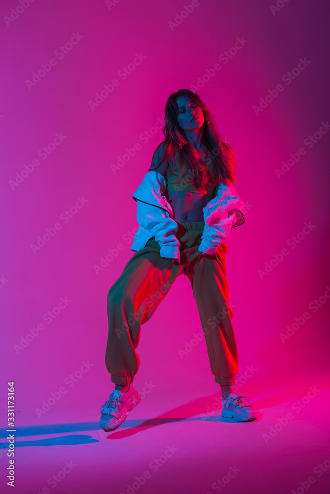 Fototapeta Fashion young woman dancer in a trendy youth tracksuit in stylish sneakers dancing hip-hop in the studio with bright pink neon color. Modern girl enjoys a dance in the studio with multi-colored light.