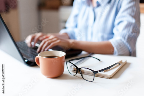 Remote working from home. Freelancer workplace in kitchen with laptop, cup of coffee, spectacles. Concept of distance learning, isolation, female business, shopping online. Close up of woman hands.