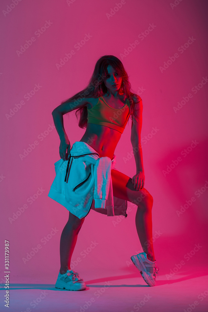 Fototapeta Cool slim young woman in fashionable top with a vintage hoodie in shoes posing with multicolored neon color background. Stylish attractive sexy girl model in the studio with bright blue disco light.