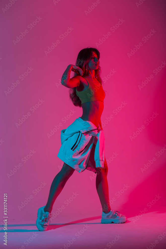 Fototapeta Fashion young woman dancer in a trendy youth clothes in stylish sneakers dancing in the studio with bright pink neon color in disco style. Girl enjoys a dance in the studio with multi-colored light.